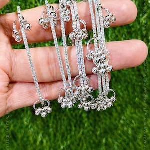 May include: Silver-toned jewelry with dangling bell-shaped charms. The jewelry is held against a green background. The text "CREATIVEGALOREIN" is visible at the bottom of the image.