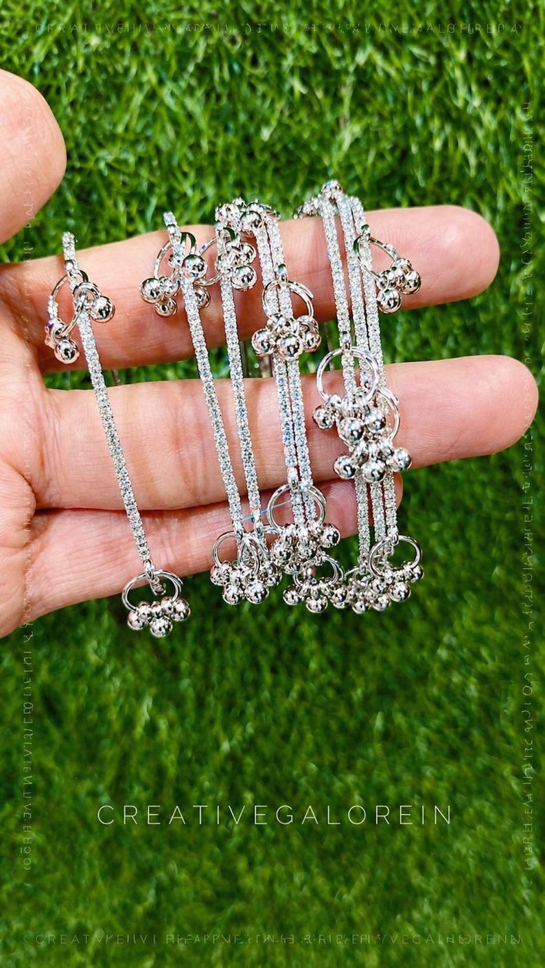 May include: Silver-toned jewelry with dangling bell-shaped charms. The jewelry is held against a green background. The text "CREATIVEGALOREIN" is visible at the bottom of the image.