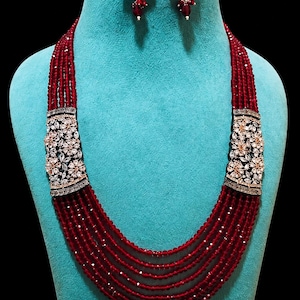 May include: A red beaded necklace and matching earrings set. The necklace features multiple strands of small red beads, with decorative floral accents. The earrings have a similar design, with red bead drops. The jewelry is displayed on a blue mannequin.