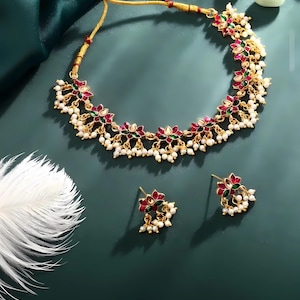 May include: A gold-coloured necklace and earring set. The necklace and earrings feature red, green, and white floral designs with small white pearl accents. The jewellery is displayed on a dark green surface.