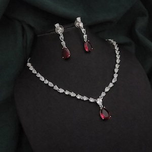 Pode incluir: Um conjunto de joias prateadas com um colar e brincos a condizer. O colar e os brincos apresentam detalhes claros e brilhantes e uma pedra preciosa vermelha escura em forma de gota. O conjunto é exibido sobre uma superfície preta.