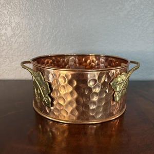 May include: A round, hammered copper bowl featuring a polished finish and two decorative brass handles shaped like oak leaves. The bowl has a wide, open top and a cylindrical body with a distinct metallic texture throughout the exterior surface.