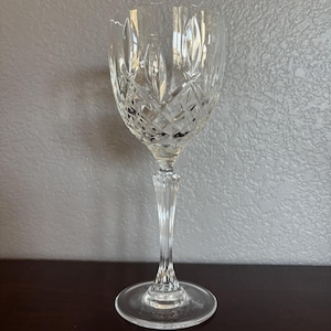 May include: A clear crystal wine glass with a diamond and leaf pattern. The glass has a long, slender stem and a round base. The glass is designed for serving wine.