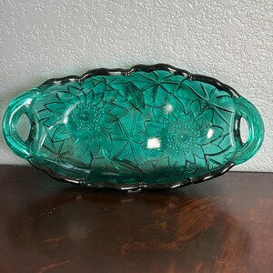 May include: A teal-colored, oval-shaped glass serving dish with a floral pattern. The dish has two handles and a scalloped edge with a dark border. The floral design is embossed on the interior of the dish.
