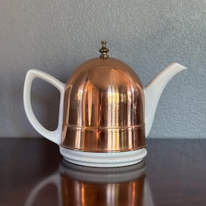 May include: A teapot featuring a white ceramic base and handle with a polished copper dome-shaped cover. The lid is topped with a small brass-colored finial. The design combines a white spout and handle with a metallic copper body and a brass knob.