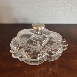 May include: A clear glass candy dish with a clover-shaped design and a silver-toned metal knob handle on the lid. The glass features etched floral and fern patterns along the top surface, with a scalloped edge and a smooth, transparent finish.