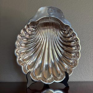 May include: A polished silver-toned metal dish shaped like a large scallop shell. The dish features deep, radiating fluted grooves that extend from the center to the scalloped edges, with a smooth, arched top section containing a small diamond cutout.