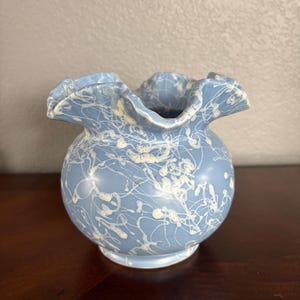 May include: A light blue ceramic vase with a round body and ruffled rim. The vase is decorated with white, splattered paint. The vase sits on a dark wooden surface. Home decor.