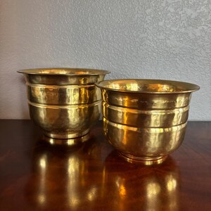 May include: Two matching round bowls crafted from polished brass, each featuring a distinct hammered texture across the exterior surface. The bowls have a wide, open top rim that tapers down to a smaller, circular base, showcasing a warm metallic gold finish.