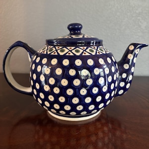 May include: A round ceramic teapot featuring a deep blue glaze decorated with a repeating pattern of white dots with dark centers. The upper section displays a geometric diamond motif in blue, white, and green, topped with a matching lid.