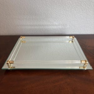 May include: A square mirrored tray featuring a clear glass border frame. The frame is accented with polished gold-tone metal corner connectors and clear glass rods that form a raised railing around the reflective surface.