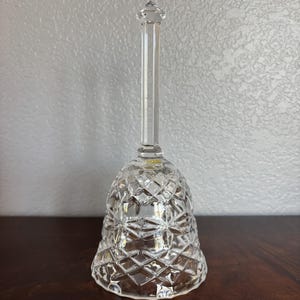 May include: A clear crystal bell with a long, slender handle and a diamond-cut pattern on the bell-shaped body. The bell has a pointed top and sits on a dark wooden surface.