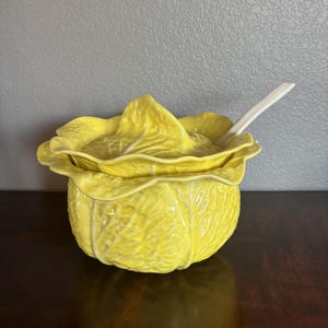 May include: A yellow ceramic cabbage-shaped serving bowl with a matching lid and a white spoon. The bowl has a textured surface and is sitting on a dark brown surface.