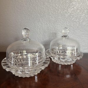 May include: Two clear glass butter dishes with matching domed lids. Each dish features a faceted, diamond-cut pattern along the base and a smooth, rounded glass cover topped with a small, vertically ribbed knob handle for easy lifting.