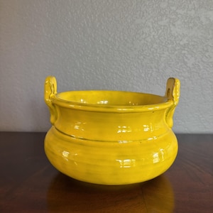 May include: A bright yellow ceramic bowl with a rounded body and two vertical handles on opposite sides. The bowl features a smooth, glossy finish with subtle variations in the yellow glaze, creating a vibrant appearance for this functional vessel.