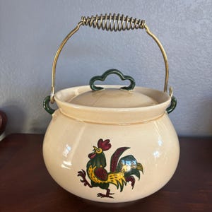 May include: Cream-colored ceramic pot with a lid and a brass handle. The pot features a colorful rooster illustration on the front. The handle has a coiled design for easy grip. The lid has a green handle. A decorative kitchen item.