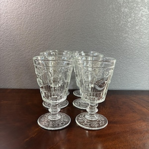 May include: A set of four clear glass goblets featuring an embossed shell and scroll pattern on the bowl, a vertical ribbed base section, a short pedestal stem, and a circular foot with a decorative etched design on the underside of the base.