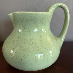 May include: A light green ceramic pitcher with a rounded body and a curved handle. The pitcher has a spout for pouring and is speckled with small brown dots. The pitcher is likely used for serving beverages.