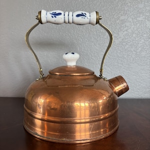 May include: A copper tea kettle with a white and blue floral patterned handle and lid knob. The kettle has a spout and a curved metal handle. The copper has a warm, reflective finish, suggesting it is well-used.