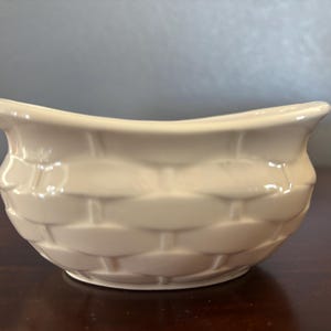 May include: A cream-colored ceramic bowl with a basket weave design. The bowl has a curved rim and sits on a dark wooden surface. The bowl is empty and appears to be new.