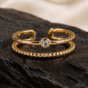 Könnte beinhalten: Ein goldfarbener Ring mit zwei Bändern und einem kleinen, klaren Edelstein. Das obere Band ist offen, während das untere Band ein strukturiertes Design aufweist. Der Ring wird auf einer dunklen, strukturierten Oberfläche mit weißem Stoffhintergrund präsentiert.