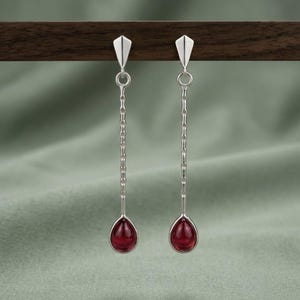 May include: A pair of silver drop earrings with a geometric design. Each earring features a teardrop-shaped, deep red gemstone at the bottom, connected by a chain to a triangular top. The earrings hang against a soft green background.