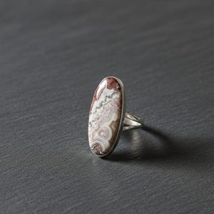 Puede incluir: Anillo de plata con una piedra ovalada que muestra un patrón distintivo de remolinos rojos, blancos y marrones. La piedra está engastada en una banda de plata pulida. El anillo es una pieza llamativa.
