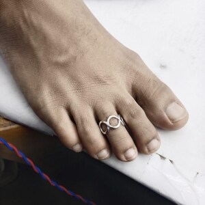 Puede incluir: Un anillo para el dedo del pie de tono plateado que presenta una serie de bucles circulares interconectados. La banda está construida con un alambre de metal continuo formado por varios círculos abiertos que envuelven el dedo del pie.