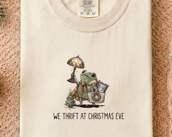 May include: A cream-colored t-shirt featuring a graphic of a green frog wearing a scarf, holding a lamp and a tote bag, with a small Christmas tree nearby. The text below the illustration reads, "WE THRIFT AT CHRISTMAS EVE."