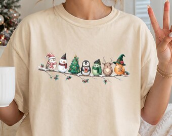 May include: A beige short-sleeve t-shirt featuring a graphic print of a row of small holiday-themed characters, including a bird, snowman, Christmas tree, penguin, and reindeer, all perched together on a thin, snow-dusted tree branch.