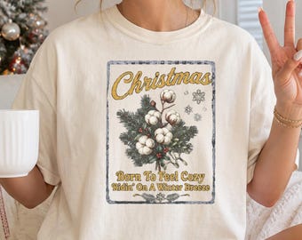 May include: A cream-colored t-shirt featuring a rectangular graphic with a central illustration of cotton bolls and pine branches. The text reads: Christmas, Born To Feel Cozy, Ridin' On A Winter Breeze, accompanied by small snowflake icons.