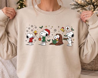 May include: A beige crewneck sweatshirt featuring a graphic print of Snoopy and Woodstock characters engaged in holiday activities, including holding a mug, reading a book, and carrying a Christmas pudding, surrounded by snowflakes and holly.