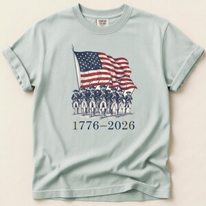 May include: Light blue t-shirt featuring an illustration of soldiers carrying the American flag. The flag is red, white, and blue. The text "1776-2026" is printed below the illustration. The shirt is made of a soft material.