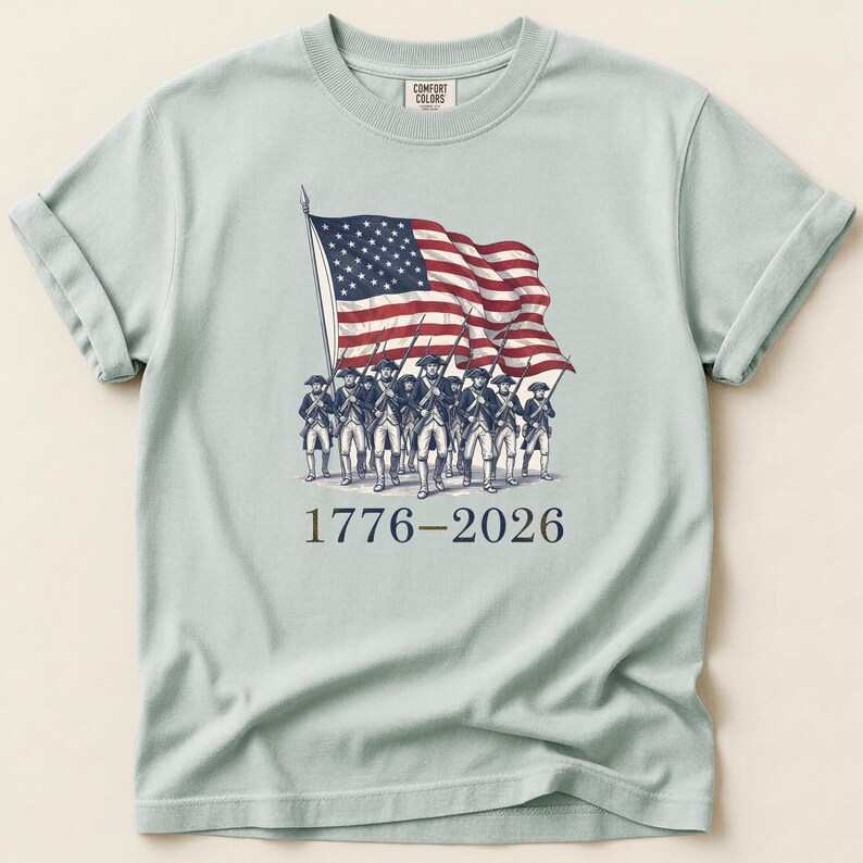 May include: Light blue t-shirt featuring an illustration of soldiers carrying the American flag. The flag is red, white, and blue. The text "1776-2026" is printed below the illustration. The shirt is made of a soft material.