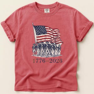 May include: Red t-shirt featuring a graphic of the American flag and soldiers. The flag is waving, and the soldiers are marching. The text "1776-2026" is printed below the graphic. The shirt has rolled-up sleeves and a crew neck.