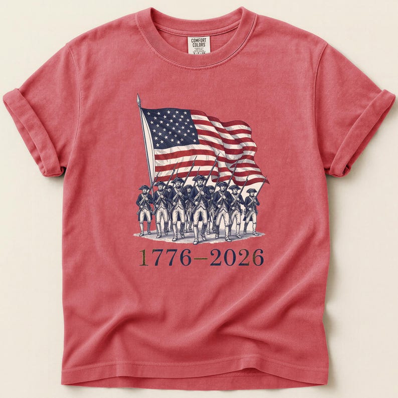 May include: Red t-shirt featuring a graphic of the American flag and soldiers. The flag is waving, and the soldiers are marching. The text "1776-2026" is printed below the graphic. The shirt has rolled-up sleeves and a crew neck.