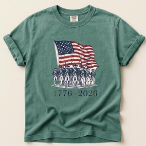 May include: A teal Comfort Colors t-shirt featuring a graphic of the American flag being carried by a group of soldiers. The text "1776-2026" is printed below the soldiers. The shirt has rolled-up sleeves and a crew neck.