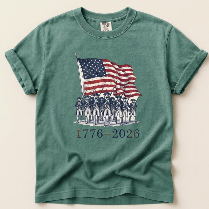 May include: A teal Comfort Colors t-shirt featuring a graphic of the American flag being carried by a group of soldiers. The text "1776-2026" is printed below the soldiers. The shirt has rolled-up sleeves and a crew neck.