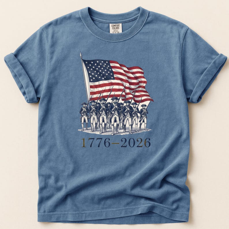 May include: Blue t-shirt featuring a graphic of the American flag and a group of soldiers marching. The text "1776-2026" is printed below the image. The shirt has rolled-up sleeves and a crew neck.
