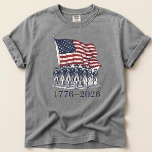 May include: Gray t-shirt featuring a vintage illustration of the American flag waving above a line of soldiers. The design includes the text "1776-2026" in a patriotic font, celebrating a historical event.