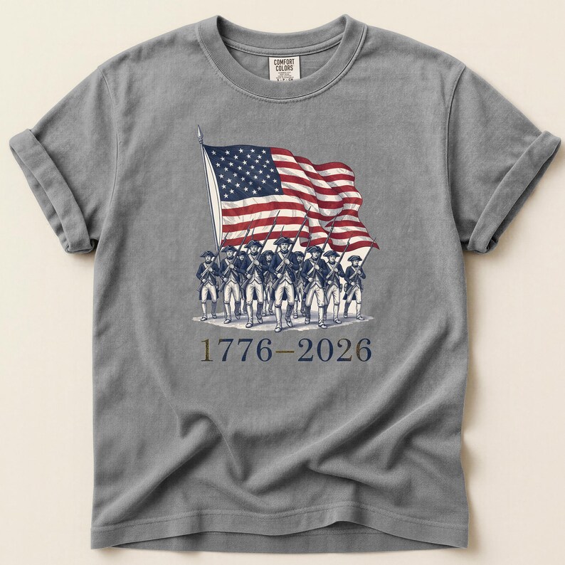 May include: Gray t-shirt featuring a vintage illustration of the American flag waving above a line of soldiers. The design includes the text "1776-2026" in a patriotic font, celebrating a historical event.