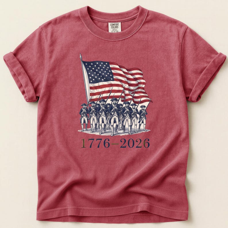 May include: Red t-shirt featuring a vintage illustration of the American flag and soldiers marching. The graphic includes the years 1776-2026. The shirt has rolled-up sleeves and a Comfort Colors tag.