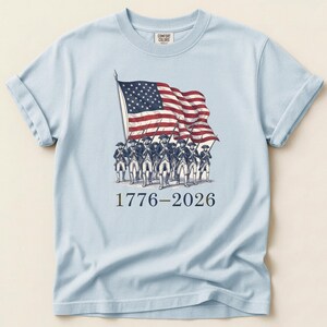 May include: Light blue t-shirt featuring a graphic of the American flag and soldiers marching. The flag is red, white, and blue. The text "1776-2026" is printed below the graphic. The shirt has rolled-up sleeves.