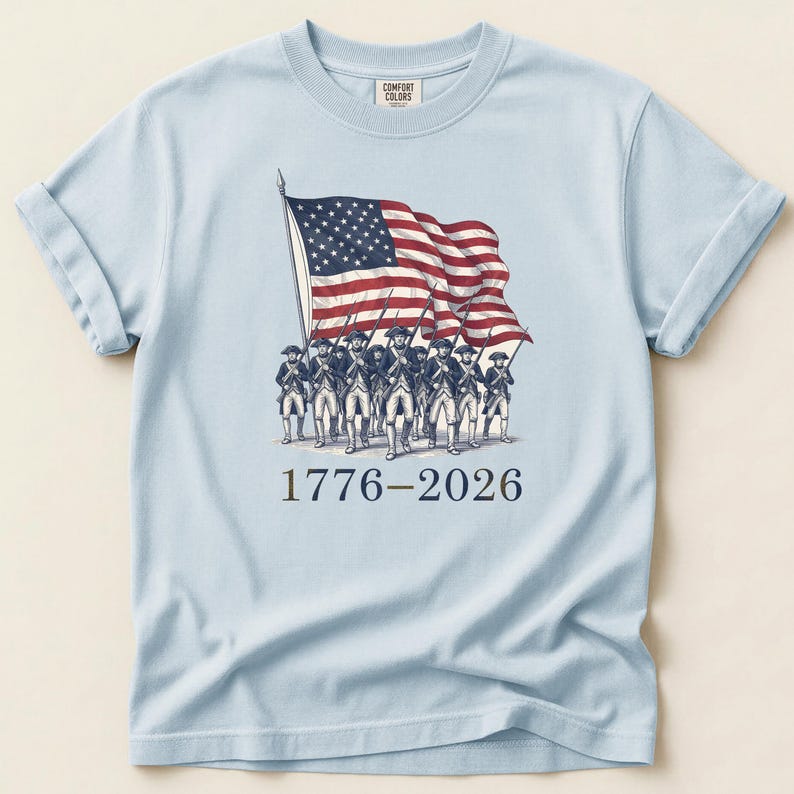 May include: Light blue t-shirt featuring a graphic of the American flag and soldiers marching. The flag is red, white, and blue. The text "1776-2026" is printed below the graphic. The shirt has rolled-up sleeves.