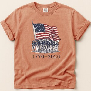 May include: A peach-colored t-shirt featuring a graphic of the American flag and soldiers. The flag is red, white, and blue, and the soldiers are in colonial uniforms. The text "1776-2026" is printed below the graphic. The shirt has rolled-up sleeves.