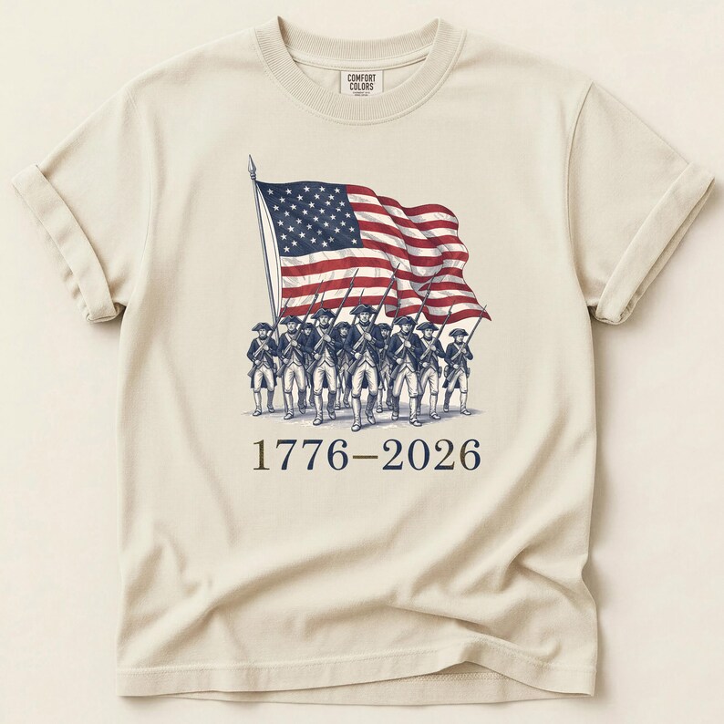 May include: Cream-colored t-shirt featuring a graphic of the American flag waving above a line of soldiers. The text "1776-2026" is printed below the image in navy blue. The shirt has rolled-up sleeves and a crew neck.