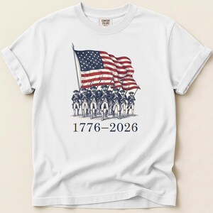 May include: White t-shirt featuring a graphic of the American flag and soldiers. The flag is red, white, and blue, and the soldiers are in colonial uniforms. The text "1776-2026" is printed below the graphic.