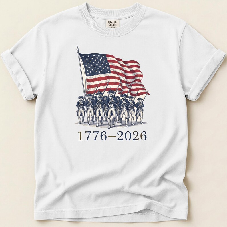 May include: White t-shirt featuring a graphic of the American flag and soldiers. The flag is red, white, and blue, and the soldiers are in colonial uniforms. The text "1776-2026" is printed below the graphic.