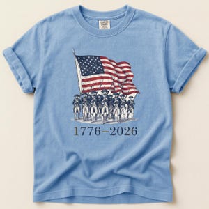 May include: Light blue t-shirt featuring a vintage illustration of the American flag and soldiers. The graphic includes the years 1776-2026. The shirt is made of a soft material and has rolled-up sleeves.