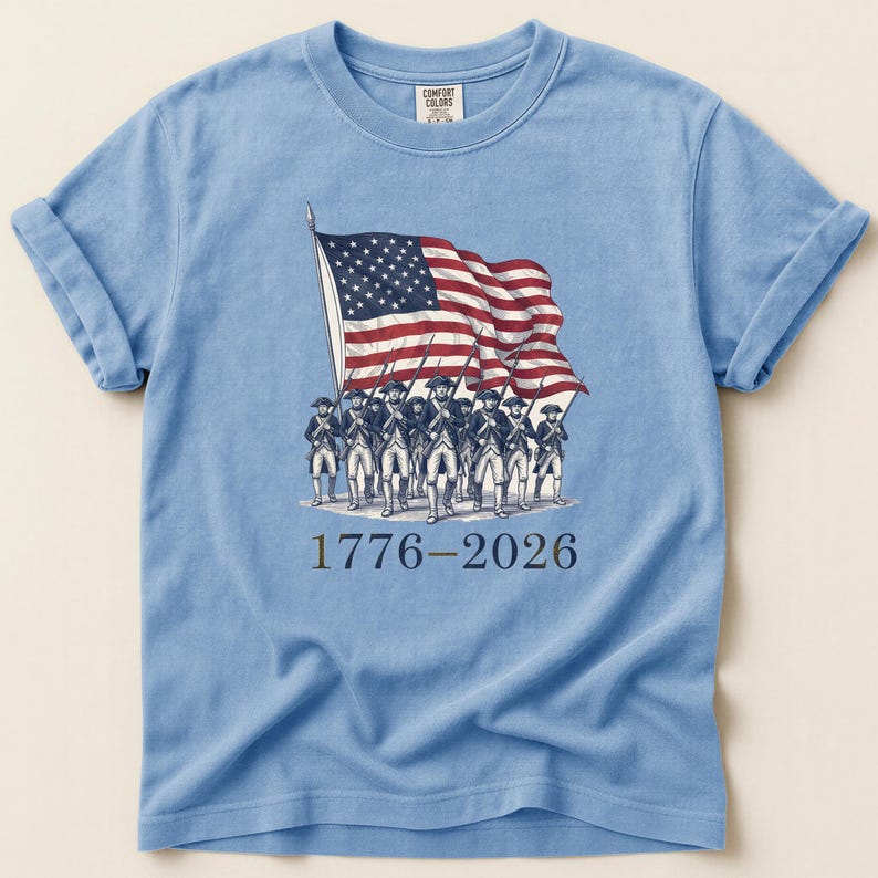 May include: Light blue t-shirt featuring a vintage illustration of the American flag and soldiers. The graphic includes the years 1776-2026. The shirt is made of a soft material and has rolled-up sleeves.